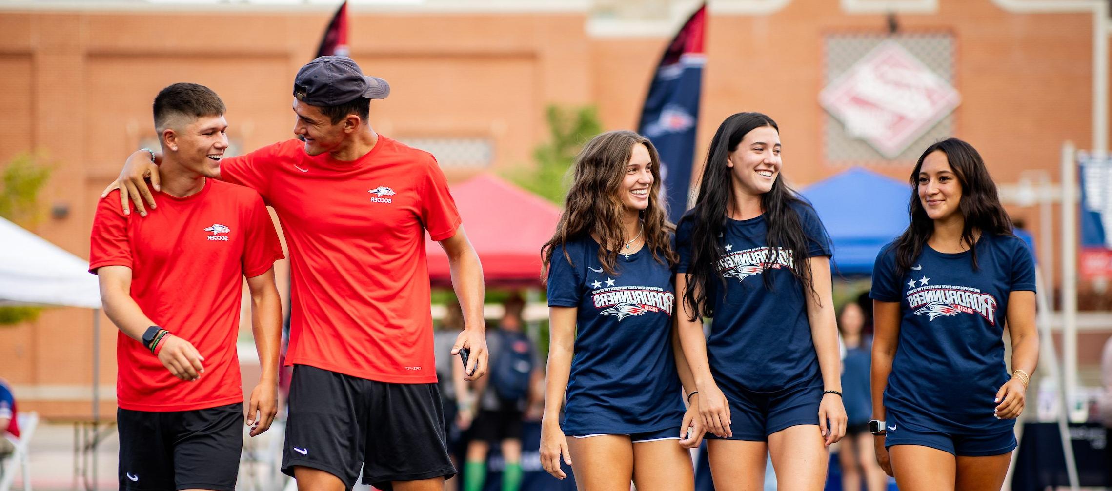 Five students walking together outside, looking at each other and smiling while wearing MSU Denver blue and red t shirts